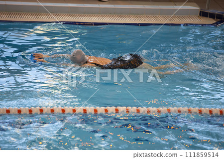 A woman swims in a black swimsuit and a swimming cap swims in the pool. 111859514