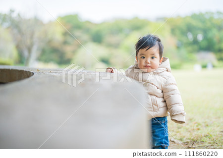 A 1-year-old toddler wearing first shoes is happily practicing tacchi with a smile in an outdoor park full of nature. A 1-year-old toddler wearing first shoes is happily practicing tacchi with a smile in an outdoor park full of nature. 111860220