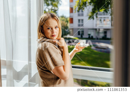 Mature blond haired woman wearing cozy sweater having calm peaceful morning standing by window with mug, drinking coffee. Elderly female enjoying herbal tea indoors. Mature blond haired woman wearing cozy sweater having calm peaceful morning standing by window with mug, drinking coffee. Elderly female enjoying herbal tea indoors. 111861353