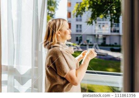 Free Time To Relax. Profile of positive 55s mature woman stand next to open window at home holding cup and drinking hot coffee. Happy lady taking break with mug near window. Free Time To Relax. Profile of positive 55s mature woman stand next to open window at home holding cup and drinking hot coffee. Happy lady taking break with mug near window. 111861356