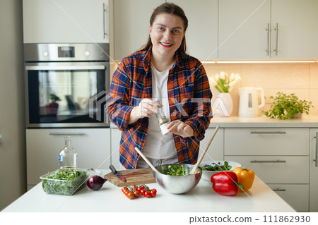 Spicing up the salad. Beautiful young woman is preparing delicious vegetable salad with tomato, arugula, red and yellow pepper in the kitchen. Healthy food. Vegan salad. Diet. Dieting concept. Healthy 111862930