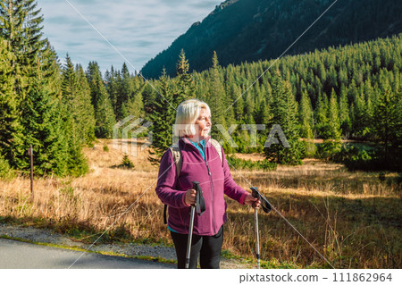 Mature caucasian blonde woman on vacation, having a hike in autumn mountains with backpack and hiking poles, enjoying adventure. Mature caucasian blonde woman on vacation, having a hike in autumn mountains with backpack and hiking poles, enjoying adventure. 111862964