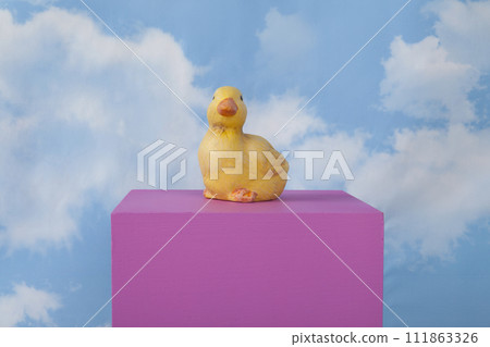 a plaster duck on a pink cube in front of a blue sky with clouds. 111863326