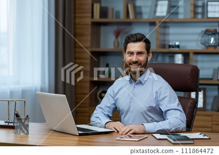 Portrait of a smiling young man wearing headphones sitting and working on a laptop in the office and looking confidently at the camera. Portrait of a smiling young man wearing headphones sitting and working on a laptop in the office and looking confidently at the camera. 111864472