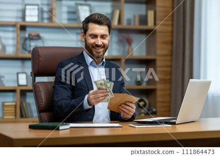 Photo of a happy young businessman sitting at a desk in the office and holding an envelope with cash in his hands, looking satisfied at the received banknotes. Photo of a happy young businessman sitting at a desk in the office and holding an envelope with cash in his hands, looking satisfied at the received banknotes. 111864473
