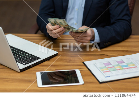 Photo concept close-up of the hand of a young man in a business suit sitting in the office at a table with gadgets, holding and counting cash dollar bills. Photo concept close-up of the hand of a young man in a business suit sitting in the office at a table with gadgets, holding and counting cash dollar bills. 111864474