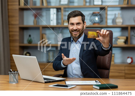 Portrait of a smiling young successful businessman, banker, financial director sitting at an office desk in a suit, holding and showing a golden plastic credit card to the camera. Portrait of a smiling young successful businessman, banker, financial director sitting at an office desk in a suit, holding and showing a golden plastic credit card to the camera. 111864475