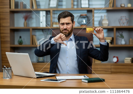 Portrait of a young businessman in a suit sitting at a desk in the office, looking at the camera, holding a credit card and pointing down with the finger of the other hand showing a negative gesture. Portrait of a young businessman in a suit sitting at a desk in the office, looking at the camera, holding a credit card and pointing down with the finger of the other hand showing a negative gesture. 111864476