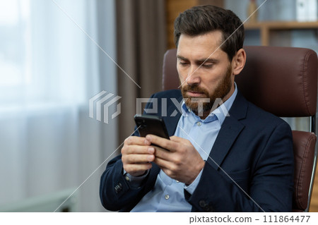 Concentrated and serious young businessman sitting in a chair in the office at the table and using a mobile phone, typing messages, texting, reading news. Concentrated and serious young businessman sitting in a chair in the office at the table and using a mobile phone, typing messages, texting, reading news. 111864477