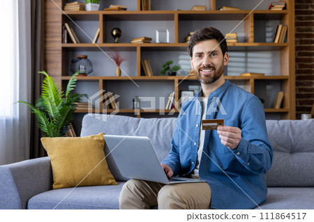 Smiling man holding a credit card and using a laptop on the couch, showcasing convenient online shopping from home. Smiling man holding a credit card and using a laptop on the couch, showcasing convenient online shopping from home. 111864517