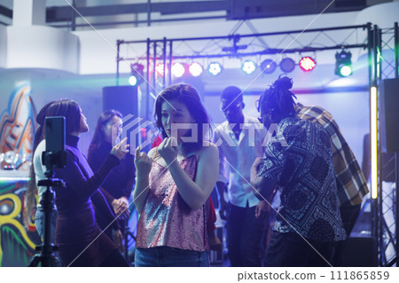 Young woman speaking on smartphone camera while recording video at nightclub party. Clubber girl having fun at club gathering and broadcasting online using mobile phone on tripod Young woman speaking on smartphone camera while recording video at nightclub party. Clubber girl having fun at club gathering and broadcasting online using mobile phone on tripod 111865859