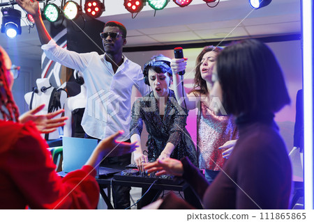 Electronic music band performing on stage in nightclub, while people partying on dancefloor. Woman dj using tracks mixer and singer giving mic to excited crowded to sing along Electronic music band performing on stage in nightclub, while people partying on dancefloor. Woman dj using tracks mixer and singer giving mic to excited crowded to sing along 111865865