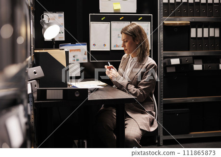 Manager sitting at desk in arhive room, checking management record trying to solve bureaucracy report. Corporate businesswoman working overtime in office, analyzing administrative documents 111865873