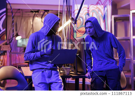 Twp hackers breaching cybersecurity system to gain illegal access. Man in hood showing computer virus code on laptop to malicious software developer and asking for advice 111867113