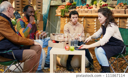 Group of cheerful people sitting at table and tasting organic products, laughing at street food fair. Customers and local vendors eating fruits and veggies at outdoors stand, produce sampling. 111867114