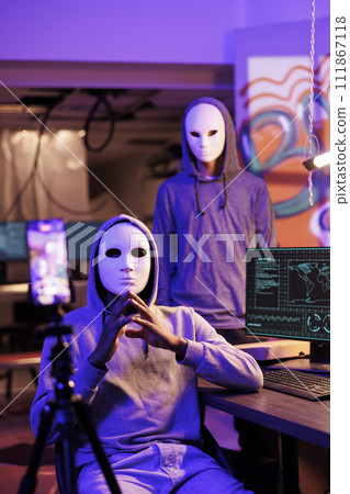 Scammers in anonymous masks streaming threat online and demand victim for payment. Hackers with hidden identity filming ransom message on mobile phone in abandoned warehouse 111867118