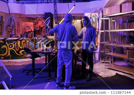 Two hackers discussing phishing attack in abandoned warehouse with graffiti. Young internet criminals in hoods planning virus malicious software programming in dark hideout workspace 111867144