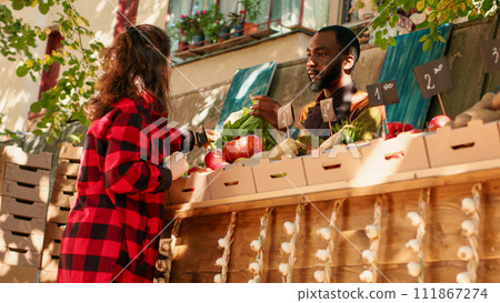 Young male seller showing natural eco produce to woman, looking to sell homegrown fruit and vegetables. Farmer presenting local healthy food at street greenmarket stand. Handheld shot. Young male seller showing natural eco produce to woman, looking to sell homegrown fruit and vegetables. Farmer presenting local healthy food at street greenmarket stand. Handheld shot. 111867274