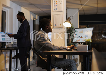 African american businessman looking at financial graphs report on computer, planning company investment typing business strategy. Executive manager working late at night in startup office African american businessman looking at financial graphs report on computer, planning company investment typing business strategy. Executive manager working late at night in startup office 111867297