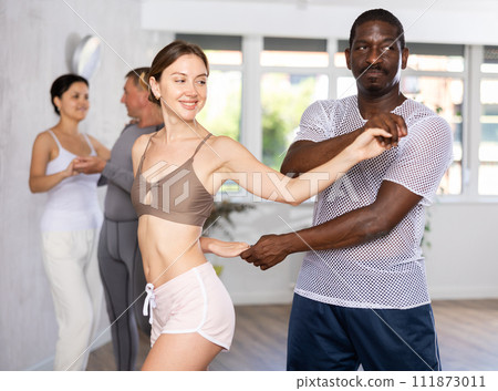 Positive African american man dancing playful Latin dance bachata with female partner during group class in choreography studio Positive African american man dancing playful Latin dance bachata with female partner during group class in choreography studio 111873011