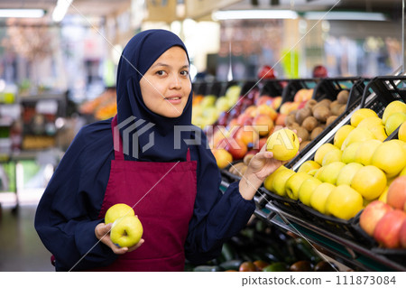Female seller in hijab and an apron puts fresh apples on shelves in the grocery section of supermarket Female seller in hijab and an apron puts fresh apples on shelves in the grocery section of supermarket 111873084