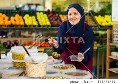 Asian female seller in hijab carefully puts large pickled olives in disposable lunch box 111873215