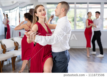Happy smiling woman dressed in red dress enjoying waltz with male partner during group party in latin dance salon. Social dancing concept Happy smiling woman dressed in red dress enjoying waltz with male partner during group party in latin dance salon. Social dancing concept 111873506
