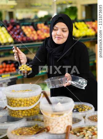 Asian woman in dark blue hijab puts handful of olives in disposable container with ladle Asian woman in dark blue hijab puts handful of olives in disposable container with ladle 111873695