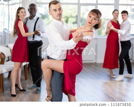 Smiling young woman wearing red dress dancing in couple rumba dance with her partner during party at dance salon Smiling young woman wearing red dress dancing in couple rumba dance with her partner during party at dance salon 111873838