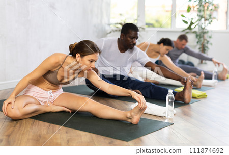 Portrait of woman making stretching legs with multinational group sitting on sports mat and making yoga exercise at fitness club 111874692