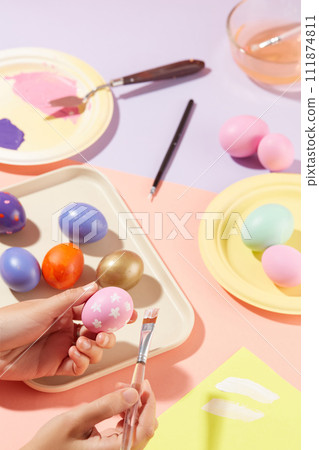 Female hand model is holding an egg in pink color and holding a paintbrush on another hand. Colorful egg are decorated on the table with a bowl of water, brushes and paint colors for Easter 111874811