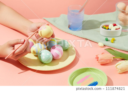Female hand model is drawing some lovely small flowers on a yellow Easter egg. A dish featured few eggs in many colors and patterns. A bowl containing candies arranged with a cup of water and brushes Female hand model is drawing some lovely small flowers on a yellow Easter egg. A dish featured few eggs in many colors and patterns. A bowl containing candies arranged with a cup of water and brushes 111874821
