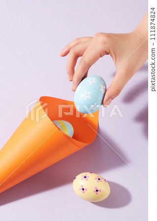 A rolled paper in orange color decorated with few Easter eggs painted with flowers and stripes. A female hand is holding a pastel blue egg. Easter Day is a special day for Christians A rolled paper in orange color decorated with few Easter eggs painted with flowers and stripes. A female hand is holding a pastel blue egg. Easter Day is a special day for Christians 111874824