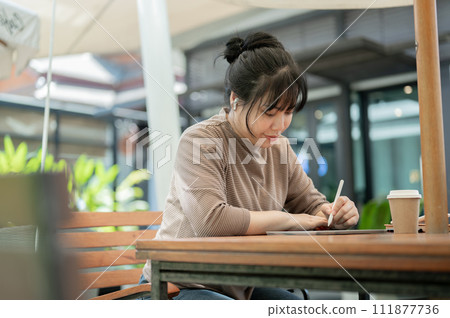 A focused Asian woman is working remotely at a cafe, wearing earbuds and using her digital tablet. A focused Asian woman is working remotely at a cafe, wearing earbuds and using her digital tablet. 111877736