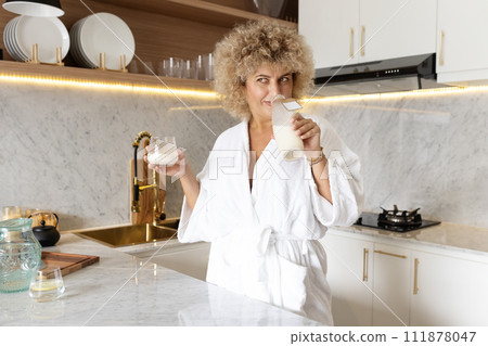 Smiling Woman in White Robe Enjoying Fresh Milk in a Modern Kitchen 111878047