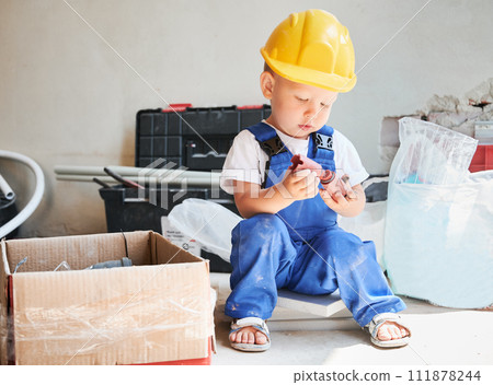 Cute little boy sitting on the floor near cardboard box with tools and materials for home renovation. Kid construction worker in safety helmet holding construction component in apartment. 111878244