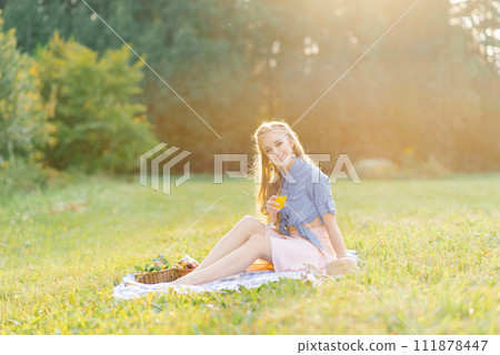 Pretty caucasian young woman with long hair is sitting on a blanket in nature on a sunny summer day. Picnic, rest, relax in the fresh air. Pretty caucasian young woman with long hair is sitting on a blanket in nature on a sunny summer day. Picnic, rest, relax in the fresh air. 111878447