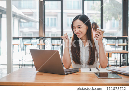 Business analytics concept, A smiling young woman enjoys her work on a laptop at a cafe table, with a coffee in hand and a bright, satisfied expression. 111878712