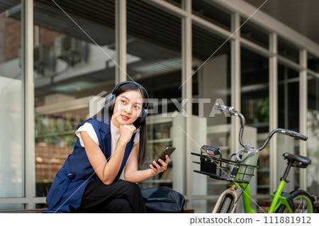 Asian businesswoman with bicycle using smartphone and sitting outside the office building. Woman commuting on bike go to work. Eco friendly vehicle, sustainable lifestyle concept Asian businesswoman with bicycle using smartphone and sitting outside the office building. Woman commuting on bike go to work. Eco friendly vehicle, sustainable lifestyle concept 111881912