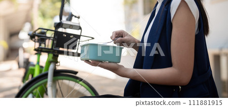 Young businesswoman while eating a lunch box sitting on a staircase during a break from work, concept healthy food using a bicycle for driving, Eco friendly vehicle, sustainable lifestyle concept Young businesswoman while eating a lunch box sitting on a staircase during a break from work, concept healthy food using a bicycle for driving, Eco friendly vehicle, sustainable lifestyle concept 111881915