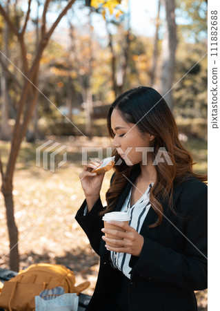 Beautiful young businesswoman with cup of coffee eating tasty sandwiches on park bench Beautiful young businesswoman with cup of coffee eating tasty sandwiches on park bench 111882688