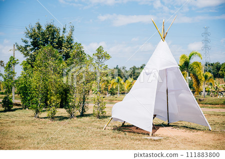 A charming summer wedding teepee stands in a green field evoking joy and celebration. The traditional wigwam adorned with dreamcatcher decorations creates a picturesque scene of family happiness. A charming summer wedding teepee stands in a green field evoking joy and celebration. The traditional wigwam adorned with dreamcatcher decorations creates a picturesque scene of family happiness. 111883800