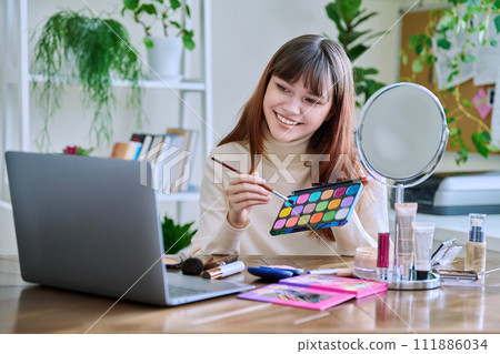 Young female showing cosmetics to laptop webcam, sitting at table at home 111886034