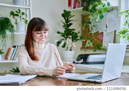 Young female college student studying at home using laptop, writing in notebook 111886036