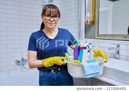 Middle-aged woman in gloves with basin of detergents in bathroom Middle-aged woman in gloves with basin of detergents in bathroom 111886488