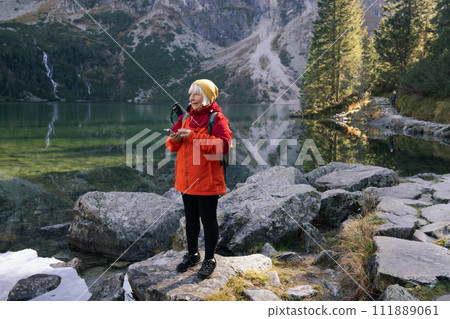 Rear shot of traveler senior woman in sportswear standing on the rocky shore and making selfie on smart phone or video. Taking photo of green hills and mountains on Morskie Oko lake. Hiking travel and Rear shot of traveler senior woman in sportswear standing on the rocky shore and making selfie on smart phone or video. Taking photo of green hills and mountains on Morskie Oko lake. Hiking travel and 111889061
