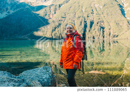 Senior 50s tourist woman with a backpack standing on top of the mountain and enjoy the beautiful view of mountains Morskie Oko lake. Hiking travel and adventure in Poland, Tatry Senior 50s tourist woman with a backpack standing on top of the mountain and enjoy the beautiful view of mountains Morskie Oko lake. Hiking travel and adventure in Poland, Tatry 111889065