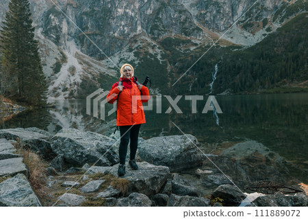 Senior 50s tourist woman with a backpack standing on top of the mountain and enjoy the beautiful view of mountains Morskie Oko lake. Hiking travel and adventure in Poland, Tatry Senior 50s tourist woman with a backpack standing on top of the mountain and enjoy the beautiful view of mountains Morskie Oko lake. Hiking travel and adventure in Poland, Tatry 111889072