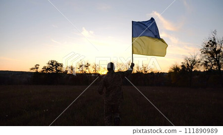 Soldier of ukrainian army running with raised blue-yellow banner on field at dusk. Young male military in uniform jogging with flag of Ukraine at meadow. Victory against russian aggression concept 111890989