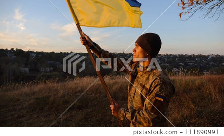 Young man in military uniform waving flag of Ukraine against beautiful sunset at background. Male ukrainian army soldier lifted national banner at countryside. Victory against russian aggression Young man in military uniform waving flag of Ukraine against beautiful sunset at background. Male ukrainian army soldier lifted national banner at countryside. Victory against russian aggression 111890991